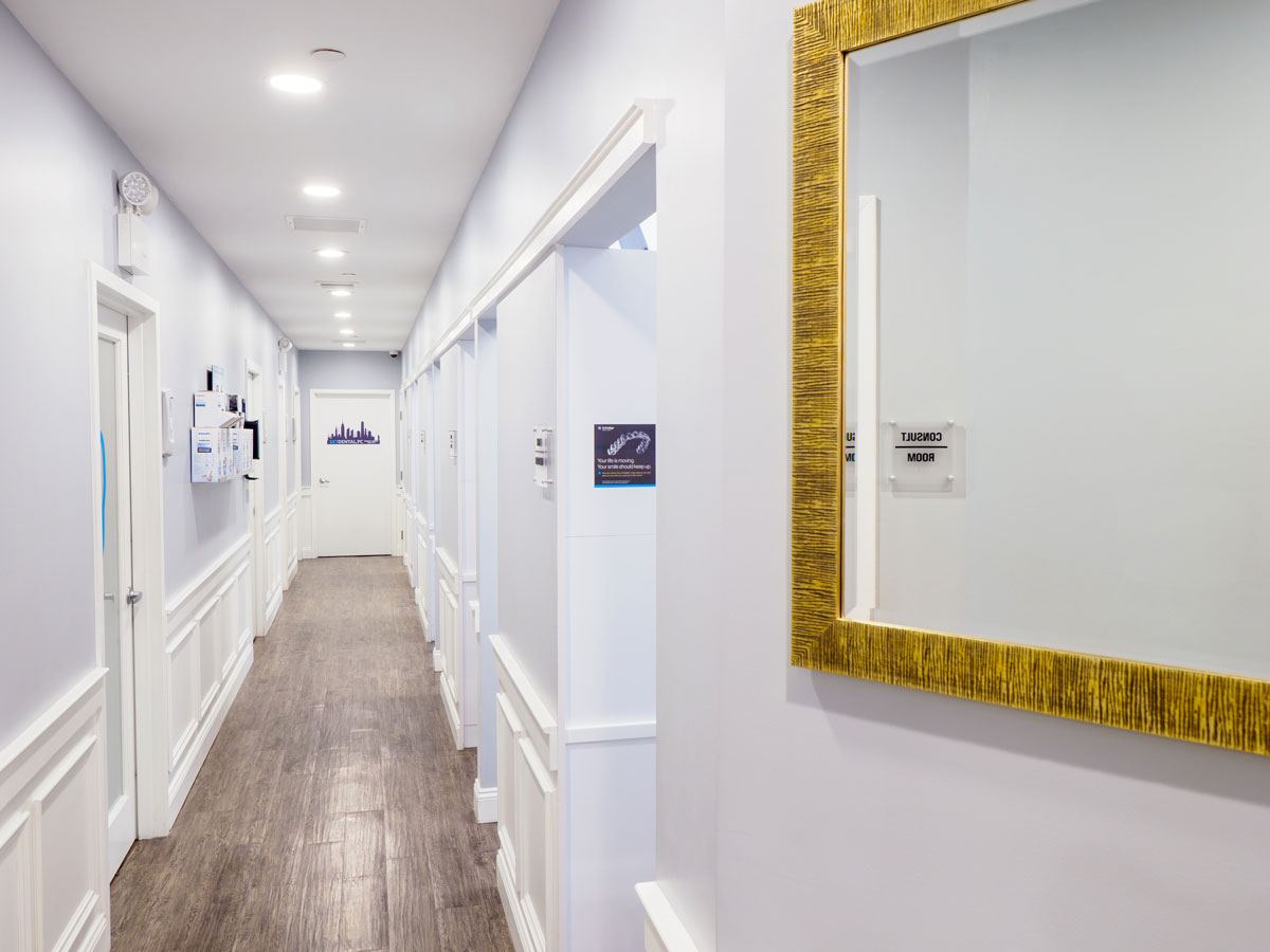 Sky Dental interior: Brightly lit, white hallway with ornate woodwork, wainscoting, and hardwood floor. Minimalist gold-framed mirror in foreground.