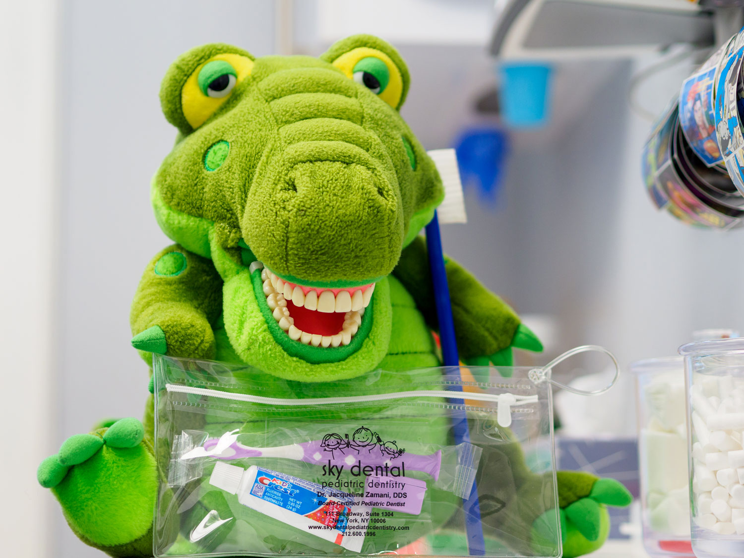 Stuffed green dinosaur with a mouthful of realistic people teeth. He holds a baggie containing a toothbrush and sample-size toothpaste.