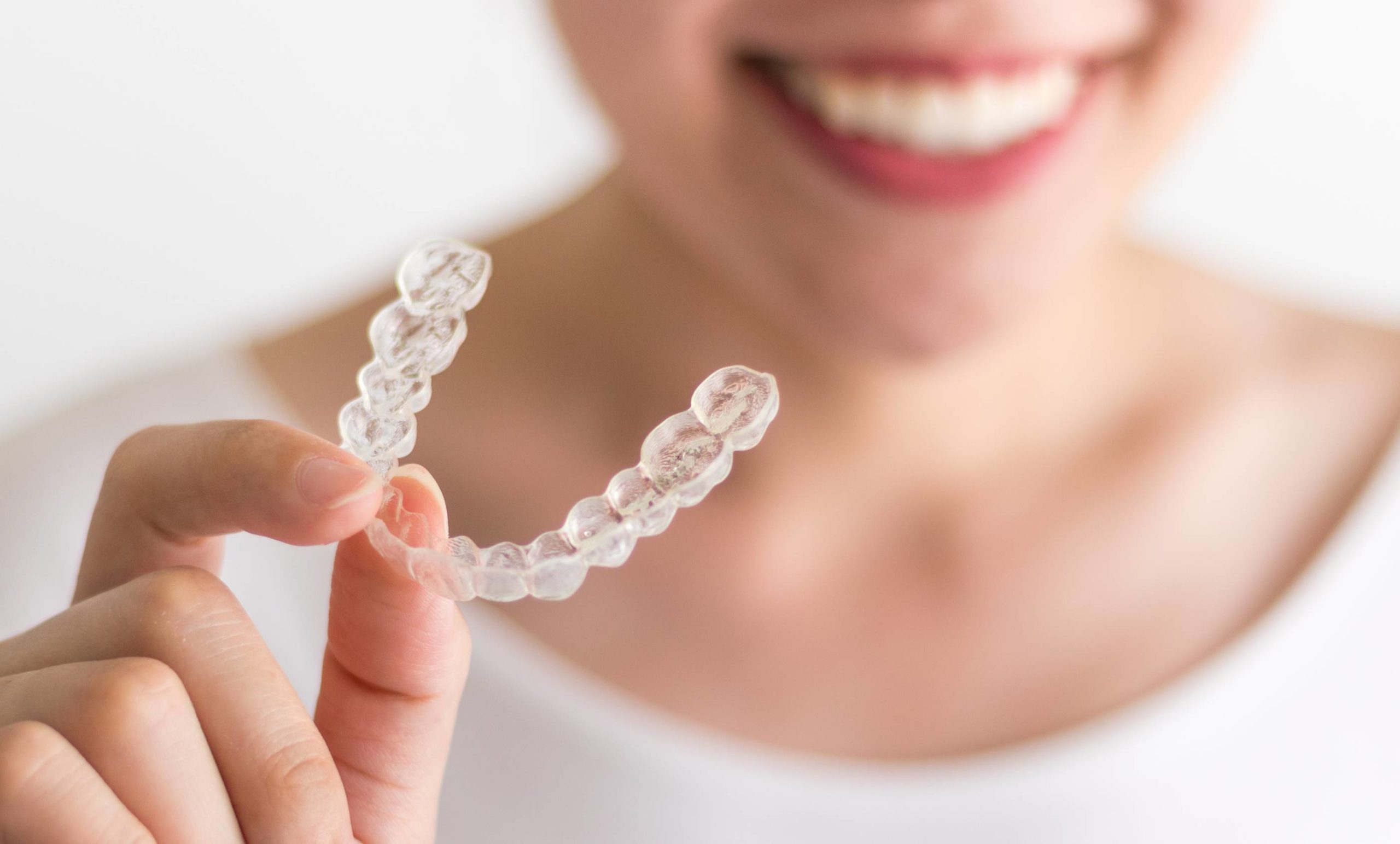 Woman with perfect teeth smiles, holding an Invisalign tray. The tray is in focus, woman's face out of focus in background.