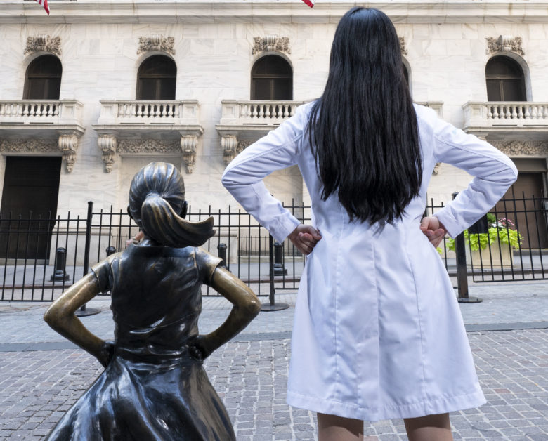 Dr. Wonnie Rhieu stands next to Fearless Girl statue facing the NY Stock Exchange. She mimics Fearless Girl's power stance.