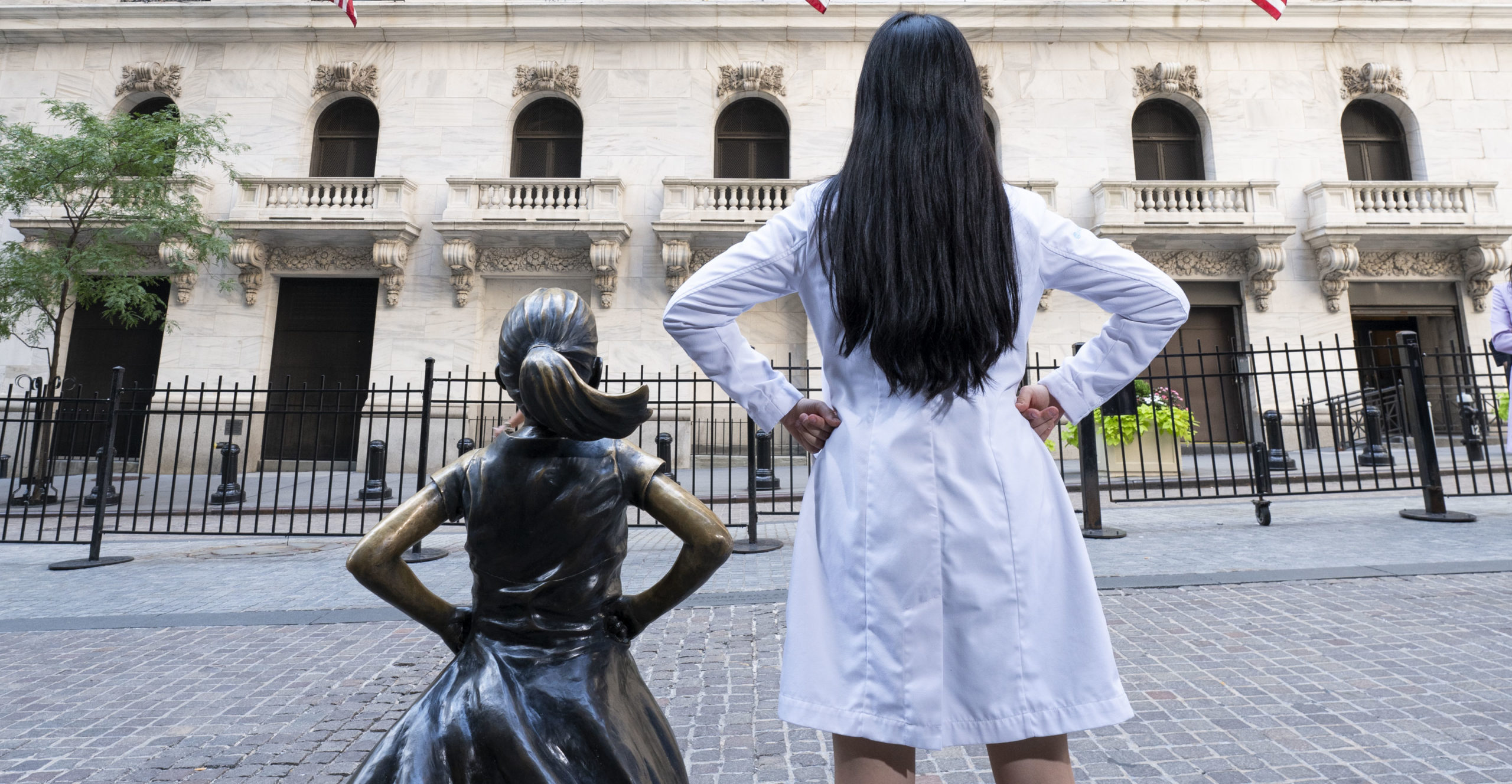 Dr. Wonnie Rhieu stands next to Fearless Girl statue facing the NY Stock Exchange. She mimics Fearless Girl's power stance.