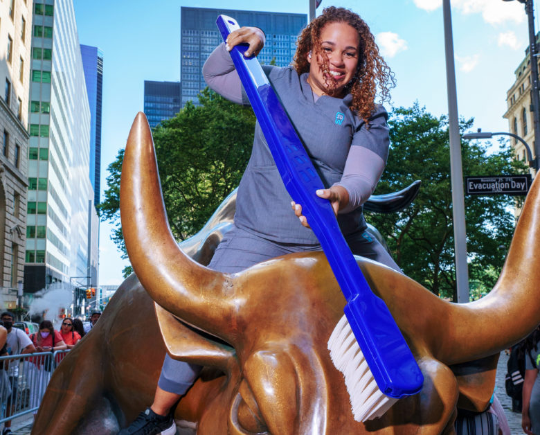 Sky Dental assistant sits smiling atop Wall Street Bull. She appears poised to brush the statue's teeth with a giant toothbrush.