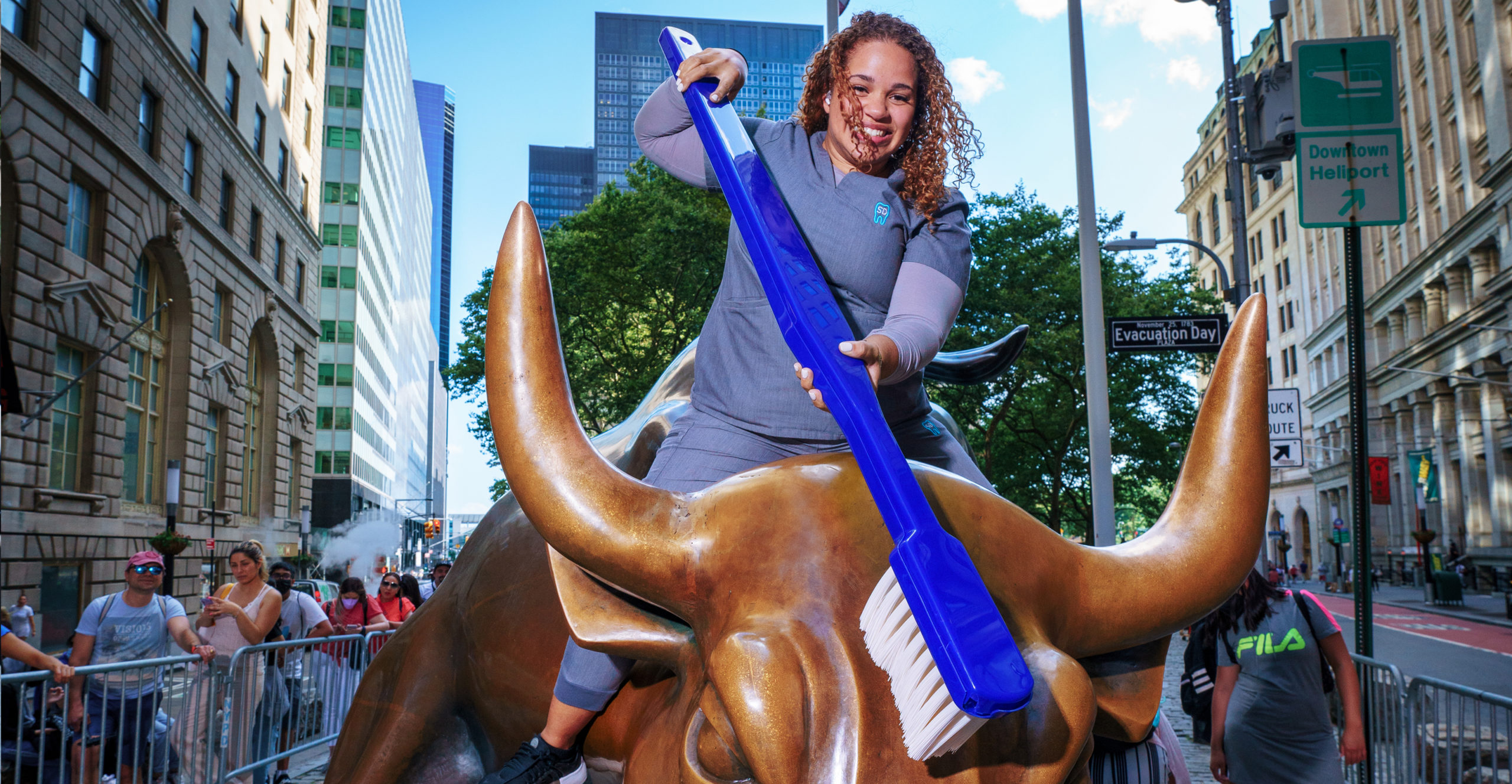 Sky Dental assistant sits smiling atop Wall Street Bull. She appears poised to brush the statue's teeth with a giant toothbrush.