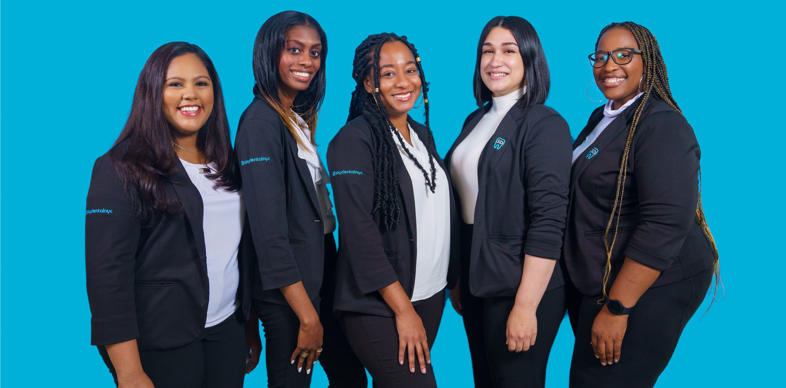 Sky Dental front desk team; a diverse group of five smiling women in white shirts and Sky Dental uniform jackets